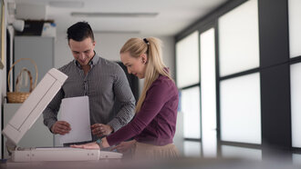 Zwei Mitarbeiter eines Unternehmens – ein dunkelhaariger Mann in schwarz-weiß-kariertem Hemd und eine blonde Frau in einem beerenfarbenen Pullover – stehen an einem von der offino Bürolösungen GmbH installierten Druck- & Scan_System mit aufgeklappter Klappe in ihren Büroräumlichkeiten