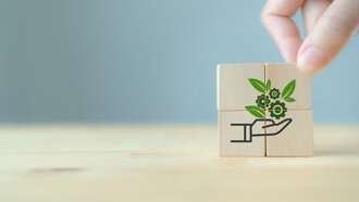 Environmental technology concept. Sustainable development goals. Konzeptionelle Darstellung der Business Ink Technology durch eine Hand, die einen Baustein auf bereits bestehende hölzerne Bausteine legt. Die Holzwürfel ergeben gemeinsam ein Icon, das technische Zahnräder mit natürlichen Blättern vereint.