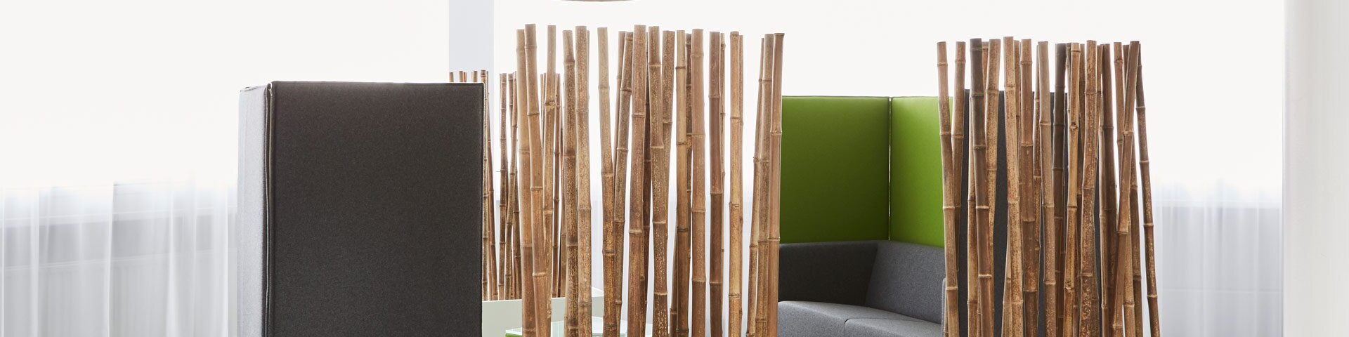 Grünes Besprechungsmobiliar nach Vorlage klassischer Alkoven mit grauem Polster im offino Showrooms in Kempten. Für den akustischen Schutz potenzieller Besprechungsteilnehmer ist das Möbelstück durch zwei Raumtrenner aus Bambus-Stangen in weißen Standfüßen von Rest des Raumes abgegrenzt.