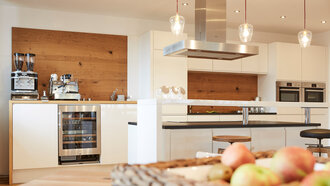 Eine Installation eines Work-Cafés im offino Showrooms in Kempten mit KAffeemaschine, Kühlschrank mit Glastür und diversen Regalen und Arbeitsflächen in eine Kombination von weißen Fronten und Echtholz-Struktur – fotografiert von der Nabe eiens Esstischs im selben Raum. Rechts im Vordergrund ist leicht unscharf eine dekorative Schale aus Korbgeflecht gefüllt mit Äpfeln zu sehen.