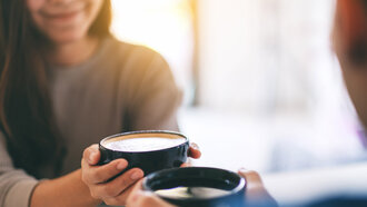 Zwei Mitarbeiter eines Unternehmens trinken gemeinsam Kaffee in einem von der offino Bürolösungen GmbH installierten Work-Café. Die Gesichter der Frau (links hinten, bildlich leicht angeschnitten) und des Mannes (rechts vorne unscharf) sind nicht zu erkennen. Der Bildfokus liegt auf den Händen der beiden, welche die schwarzen, gut gefüllten Tassen mit Milchschaumgetränken halten.