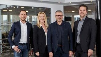 Das Führungsteam der offino Bürolösungen GmbH – namentlich Fabio Leßwing, Isabella Leßwing, Horst Leßwing und Jörg Wegmann (v.l.n.r.) – posiert in dunkler Geschäftskleidung und sympathisch lächelnd vor dem Treppenabgang im ersten Stock des offino Showrooms in Kempten