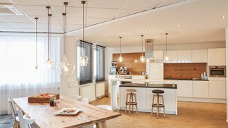 Perpektivische Totale einer Work-Kitchen im Showroom der offino Bürolösungen GmbH in Kempten. Zu sehen ist eine Küchenzeile (Materialmix aus weißen Hochglanz-Fronten und Echtholz) mit Kaffeemaschinen, Spülbecken, diversen Schränken und einer Mikrowelle. Mittig ist eine Kücheninsel mit Auflage-Bar und Sitzmöglichkeiten auf Barstühlen positioniert. Links im Raum ist ein Massivholztisch mit unetrschiedlichen weißen Stühlen zu sehen. Auf dem Tisch ist neben einem zubereiteten Pasta-Gericht und diversen Fruchsaftflaschen und Trinkgläsern auch eine Schale aus Korbgeflecht mit frischen Äpfeln zu sehen.