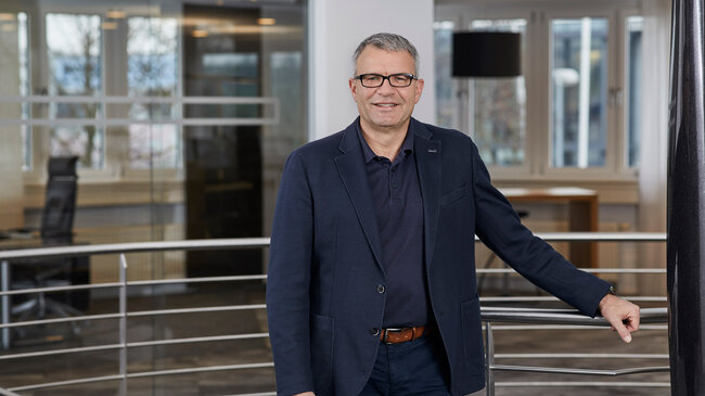 Horst Leßwing Horst Leßwing – Mitglied im Führungsteam der offino Bürolösungen GmbH – posiert in dunkler Geschäftskleidung und sympathisch lächelnd vor dem Treppenabgang im ersten Stock des offino Showrooms in Kempten